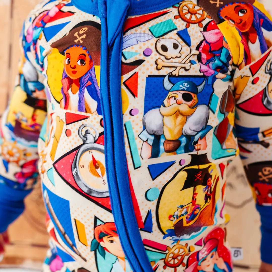 Close-up of a colorful pirate-themed onesie fabric with a blue zipper, showing cartoon pirate characters, a skull and crossbones, a compass, and a ship